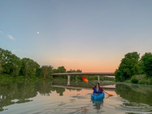 A woman kayaks on the Grand River at dusk. The bridge and the moon are seen just ahead of her.